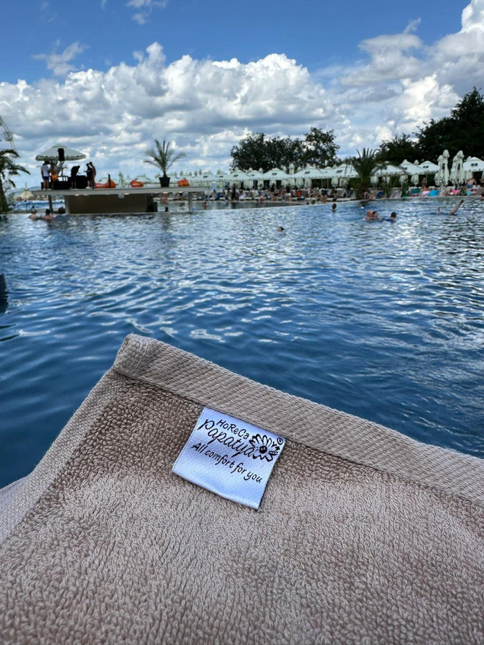 Prosop bej HoReCa Papatya cu etichetă brodată, ținut în prim-plan lângă piscina exterioară cu scenă și umbrele aliniate.