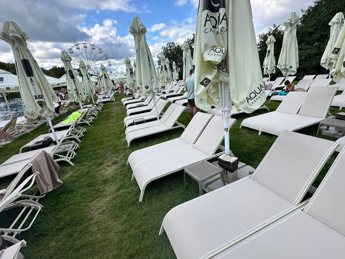 Rânduri de șezlonguri și umbrele închise pe iarbă, cu roata panoramică și piscina exterioară vizibile în depărtare.