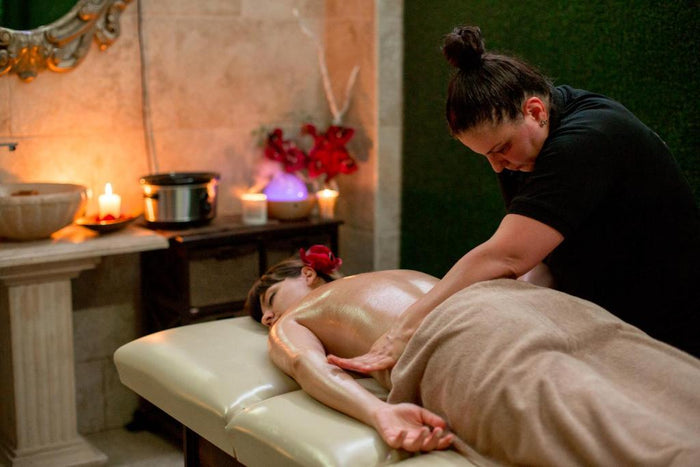 Terapeut aplicând un masaj cu ulei pe un pat SPA, într-o cameră decorată cu lumânări aprinse și flori roșii.